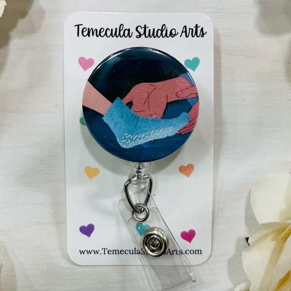 May include: A circular badge reel featuring a graphic illustration of a hand placing a light blue sock onto a foot against a dark blue background. The badge is mounted on a white display card with the text Temecula Studio Arts and small hearts.