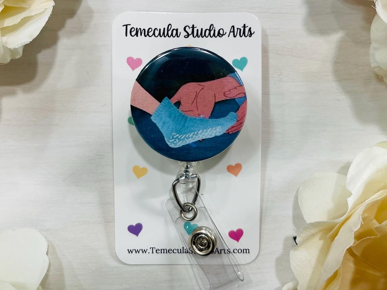May include: A circular badge reel featuring a graphic illustration of a hand placing a light blue sock onto a foot against a dark blue background. The badge is mounted on a white display card with the text Temecula Studio Arts and small hearts.