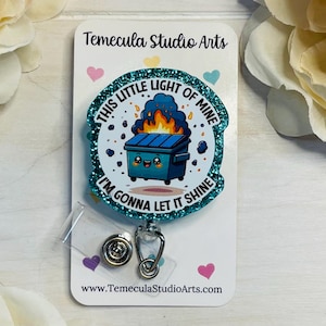 May include: A teal blue glitter badge holder with a cartoon image of a trash can with a smiling face and flames coming out of it. The text on the badge holder reads "This little light of mine, I'm gonna let it shine."