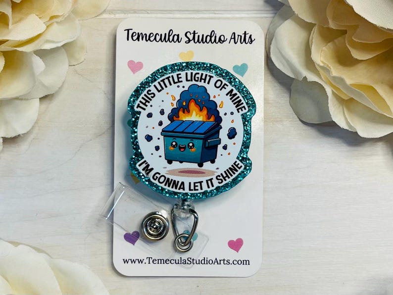 May include: A teal blue glitter badge holder with a cartoon image of a trash can with a smiling face and flames coming out of it. The text on the badge holder reads "This little light of mine, I'm gonna let it shine."