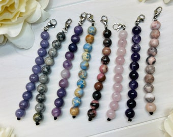 Puede incluir: Siete pulseras de cuentas con cierres plateados. Las pulseras están hechas con piedras de diferentes colores, incluyendo morado, rosa, marrón y gris.