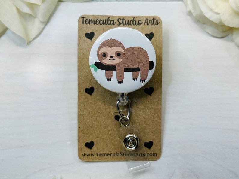 May include: A white button with a brown sloth hanging from a branch. The button is attached to a retractable badge holder with a silver clip. The background is a brown card with the text "Temecula Studio Arts" and "www.TemeculaStudioArts.com".