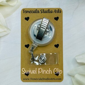 May include: A clear retractable badge holder with a silver swivel pinch clip. The holder is on a brown card with black hearts and the text "Temecula Studio Arts" and "Swivel Pinch Clip" with the website "www.TemeculaStudioArts.com".