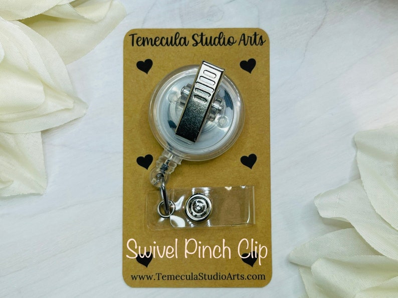 May include: A clear retractable badge holder with a silver swivel pinch clip. The holder is on a brown card with black hearts and the text "Temecula Studio Arts" and "Swivel Pinch Clip" with the website "www.TemeculaStudioArts.com".