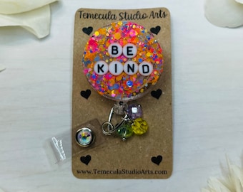 May include: A round, clear badge holder with a colorful glitter background and the words "BE KIND" in white letters. The badge holder is attached to a silver chain with colorful beads.