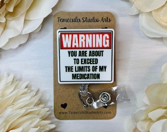May include: A white and red warning sign with the text "WARNING YOU ARE ABOUT TO EXCEED THE LIMITS OF MY MEDICATION" on a brown card with a silver retractable badge holder.