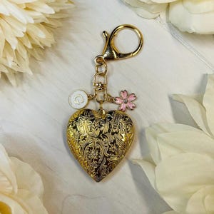 May include: Gold heart-shaped locket keychain with a floral design, a small charm with the letter "G" and a pink flower charm.