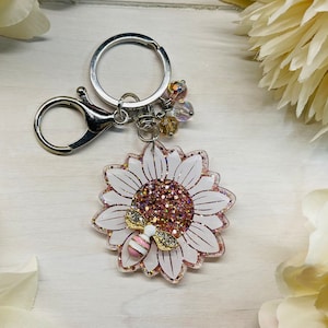 May include: A white and gold glitter sunflower keychain with a small bee charm. The keychain has a silver split ring and a silver lobster clasp.