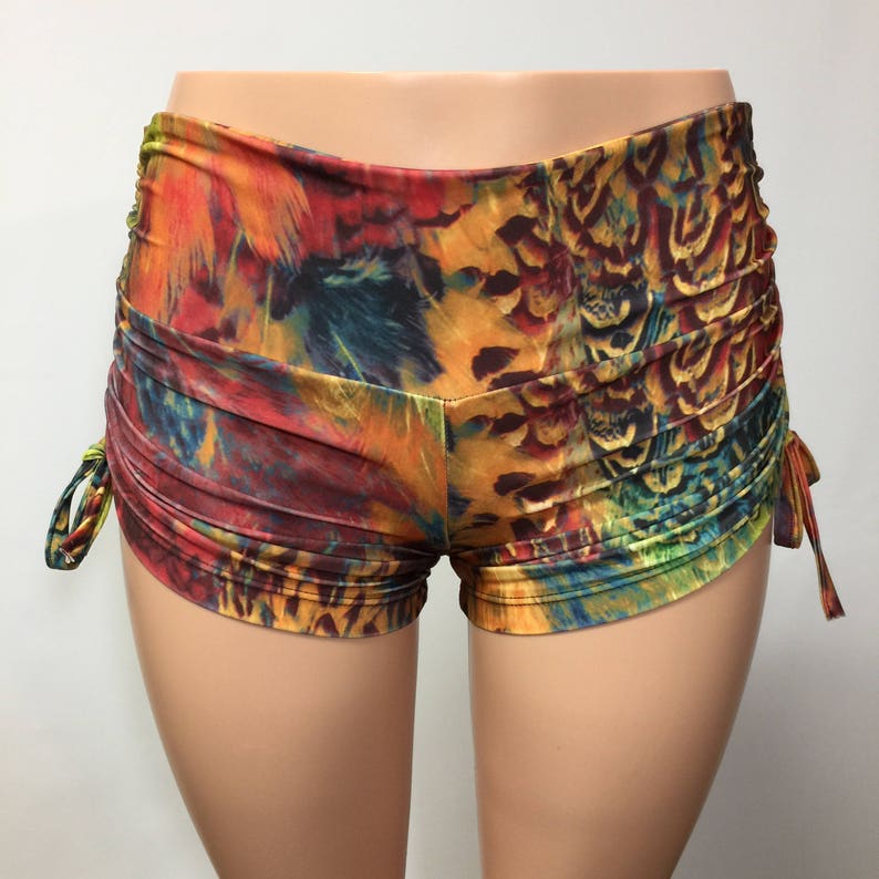 May include: High-waisted shorts featuring a vibrant, multicolored feather print in shades of orange, yellow, blue, and red. The garment includes ruched side panels with adjustable drawstring ties on both sides, creating a gathered texture.