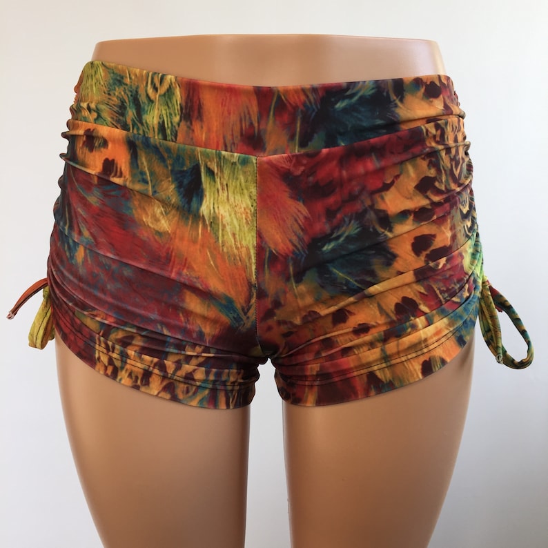 May include: High-waisted shorts featuring a vibrant, multi-colored feather print in shades of orange, red, yellow, blue, and black. The shorts include ruched side panels with adjustable drawstring ties on both sides, creating a gathered texture.