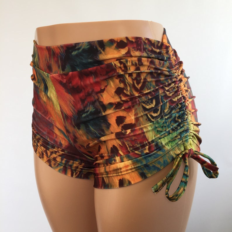 May include: High-waisted shorts featuring a vibrant, multi-colored abstract print in shades of orange, red, yellow, green, and black. The garment includes a side drawstring detail that creates gathered, ruched fabric along the right hip area.
