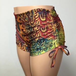 May include: High-waisted shorts featuring a vibrant, multicolored feather-inspired print in shades of red, orange, yellow, green, and blue. The garment includes side ruching details with adjustable fabric ties that create a gathered effect.