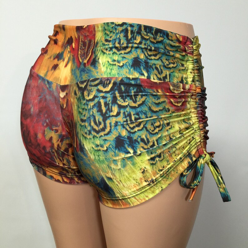 May include: High-waisted shorts featuring a vibrant, multicolored peacock feather print in shades of green, blue, yellow, and red. The garment includes a side drawstring detail with adjustable ties that create a gathered, ruched effect along the hip.