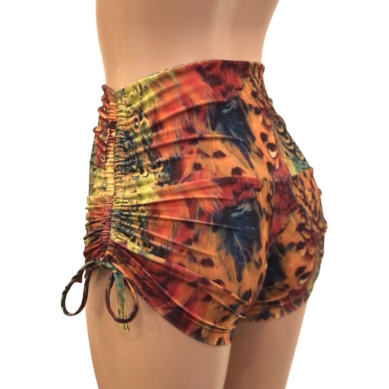 May include: High-waisted shorts featuring a vibrant, multi-colored abstract print with shades of orange, yellow, blue, and black. The garment includes side ruching details with adjustable drawstring ties that create a gathered effect along the hips.