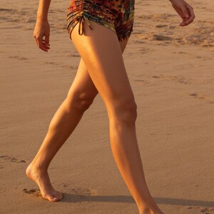 May include: High-waisted shorts featuring a multi-colored abstract print in shades of orange, yellow, and dark brown. The shorts include adjustable side drawstrings with ties that create a gathered, ruched effect along the outer leg seams.