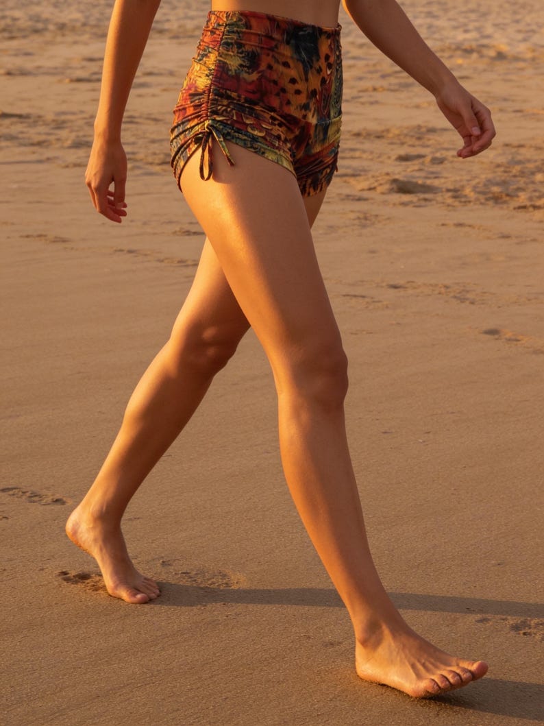 May include: High-waisted shorts featuring a multi-colored abstract print in shades of orange, yellow, and dark brown. The shorts include adjustable side drawstrings with ties that create a gathered, ruched effect along the outer leg seams.