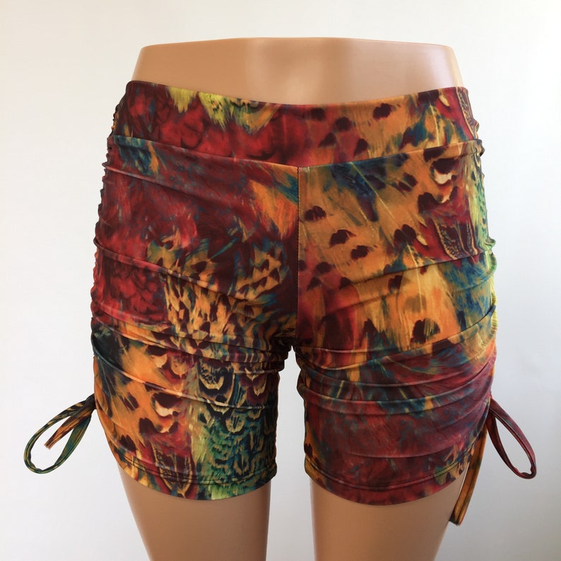 May include: Shorts featuring a vibrant, multicolored feather print in shades of red, orange, yellow, and green. The garment has a wide waistband and adjustable side ties that create a ruched effect along the outer seams of the legs.