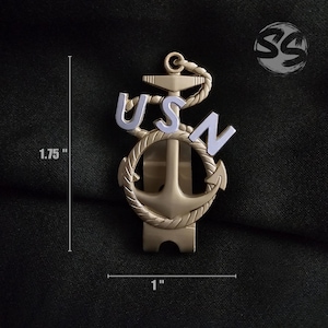 May include: A gold-colored USN anchor pin, approximately 1.75 inches tall and 1 inch wide. The pin features the letters "USN" in light blue, with an anchor design and rope detailing. The pin is set against a dark background.