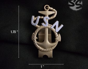 May include: A gold-colored USN anchor pin, approximately 1.75 inches tall and 1 inch wide. The pin features the letters "USN" in light blue, with an anchor design and rope detailing. The pin is set against a dark background.