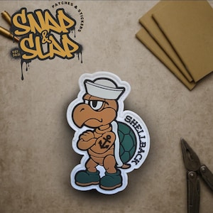 Peut inclure: Un écusson de dessin animé représentant une tortue portant un chapeau de marin, les bras croisés et un tatouage d'ancre. L'écusson est bordé de blanc avec le texte "SHELLBACK". Le coin supérieur gauche porte le texte "SNAP & SLAP" et "PATCHES & STICKERS".