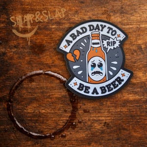 May include: A circular patch featuring a cartoon orange beer bottle with a sad, crying face. The text reads A BAD DAY TO RIP and BE A BEER. The design includes a broken heart icon and small star accents, rendered in black, white, and orange colors.