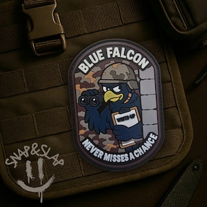 May include: A rectangular morale patch featuring a cartoon bird wearing a camouflage helmet, holding binoculars and a clipboard labelled WRITE-UP. The text BLUE FALCON and NEVER MISSES A CHANCE surrounds the bird against a camouflage background.