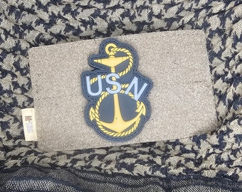 May include: A yellow and black anchor with a white "USN" in the center. The anchor is on a brown background.