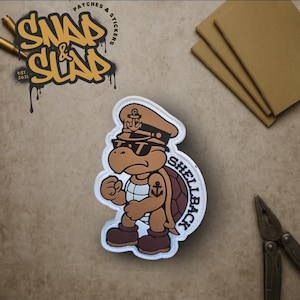 Peut inclure: Un écusson de tortue de dessin animé portant un chapeau de capitaine et des lunettes de soleil, avec le mot "SHELLBACK" sur le côté. L'écusson est marron, beige et blanc, avec une ancre sur le chapeau et la poitrine. L'écusson est sur une surface beige avec les mots "SNAP & SLAP" en jaune.