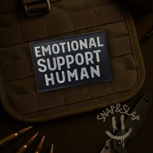 May include: A rectangular black morale patch featuring the white text EMOTIONAL SUPPORT HUMAN in a bold sans-serif font. The patch is attached to a dark olive green tactical gear panel with horizontal webbing straps and a plastic buckle.