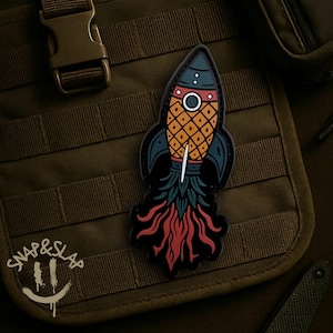 May include: A colorful patch featuring a rocket ship design. The rocket has a pineapple-like body in orange and yellow, with blue and black accents. The rocket's exhaust is red and green. The patch has the text "SNAP & SLAP" and a smiley face.