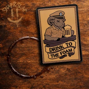 May include: A rectangular patch with a cartoon ram in a naval uniform holding a beer mug. The patch has a tan background with a black border. The text "DRINK TO THE FOAM" is on a banner below the ram.