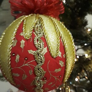 Może przedstawiać: Czerwona ozdoba choinkowa ze złotymi akcentami i dekoracyjnym złotym wykończeniem. Ozdoba ma złote liście ostrokrzewu i złotą wstążkę. Wisi na choince.