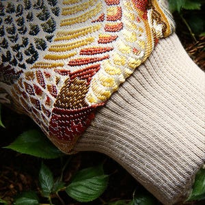 May include: A close-up of a beige knitted fabric with a colorful patterned trim. The fabric is textured and has a ribbed appearance. The trim features a geometric design with shades of red, yellow, green, and blue.