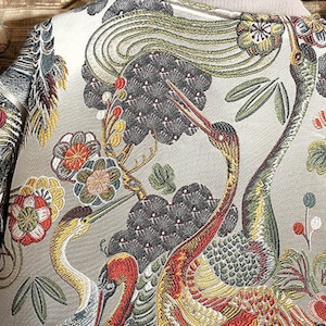 May include: A beige fabric with an embroidered design of cranes and flowers. The cranes are in shades of yellow, red, and green, and the flowers are in shades of red, pink, and white.