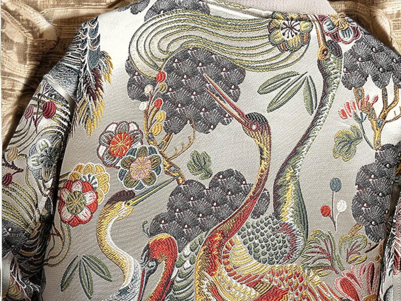 May include: A beige fabric with an embroidered design of cranes and flowers. The cranes are in shades of yellow, red, and green, and the flowers are in shades of red, pink, and white.