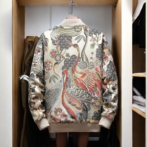 May include: A cream-colored bomber jacket with a floral and bird print. The print features red, orange, yellow, and black colors. The jacket has a zipper closure and long sleeves.