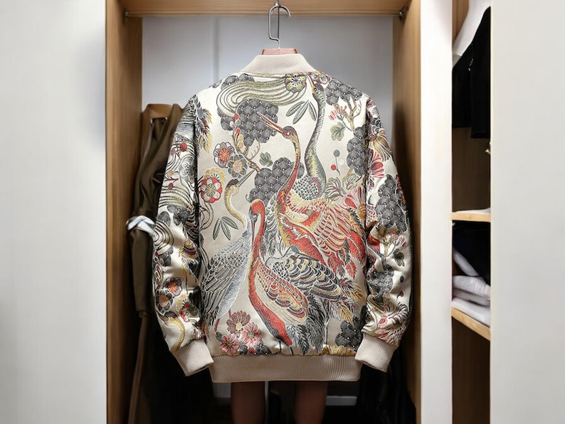 May include: A cream-colored bomber jacket with a floral and bird print. The print features red, orange, yellow, and black colors. The jacket has a zipper closure and long sleeves.