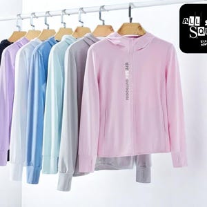 Puede incluir: Una fila de camisas con cremallera y capucha de manga larga en varios colores pastel, incluyendo negro, lavanda, azul claro y rosa, colgadas en una percha metálica. La camisa rosa tiene el texto "UPF 50+ OUTDOORS" impreso. Un cuadrado negro con el texto "ALL STYLE SQUAD" está en la esquina superior derecha.