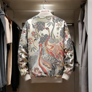 May include: A cream-colored bomber jacket with a colorful floral and bird print. The print features cranes, flowers, and leaves in shades of red, orange, yellow, green, and blue.