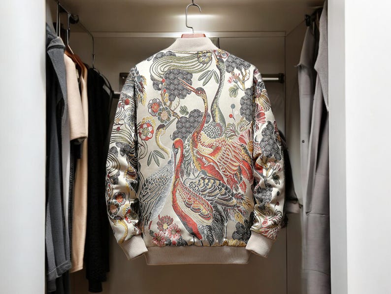 May include: A cream-colored bomber jacket with a colorful floral and bird print. The print features cranes, flowers, and leaves in shades of red, orange, yellow, green, and blue.