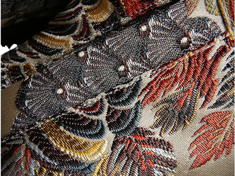 May include: Close-up of a fabric with an intricate embroidered design. The fabric features a variety of colors, including gold, red, brown, and blue. The design is made up of leaves and flowers.