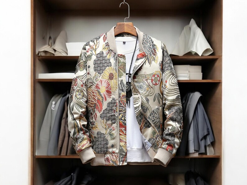 May include: A beige bomber jacket with a floral print featuring red, yellow, and blue flowers. The jacket has a zipper closure and a ribbed collar, cuffs, and hem.