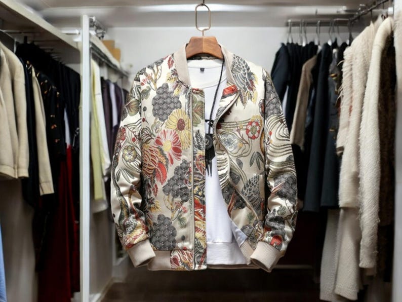 May include: A beige bomber jacket with a floral print featuring red, yellow, and green flowers. The jacket has a zipper closure and a ribbed collar, cuffs, and hem.