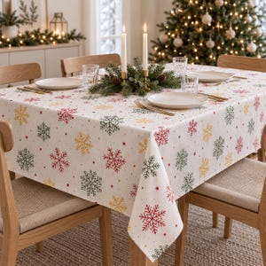 Könnte beinhalten: Eine quadratische weiße Tischdecke mit einem wiederkehrenden Muster aus roten, grünen und goldenen Schneeflocken sowie kleinen Sternen. Der Stoff liegt auf einem Esstisch, der mit weißen Tellern, klaren Gläsern und goldenem Besteck gedeckt ist.