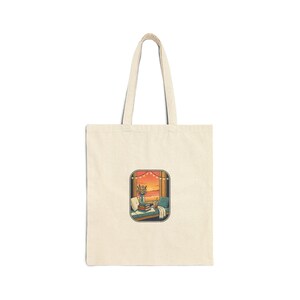 May include: Cream-colored tote bag featuring a rectangular illustration of a cozy reading nook with a sunset view. The illustration includes books, a cup, and a small plant. The bag has a simple design with long handles.
