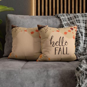 Può includere: Due cuscini quadrati con sfondo beige e design autunnale. I cuscini presentano foglie arancioni e rosse, con le parole "hello FALL" in un carattere scritto a mano. Sono appoggiati su un divano grigio.