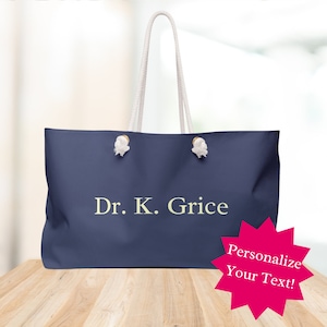 Puede incluir: Una bolsa de lona azul marino con asas de cuerda blanca y el texto "Dr. K. Grice" en una fuente amarilla clara. La bolsa está sentada sobre una superficie de madera. Un estallido de estrella rosa con el texto "¡Personaliza tu texto!" está en la esquina inferior derecha de la imagen.