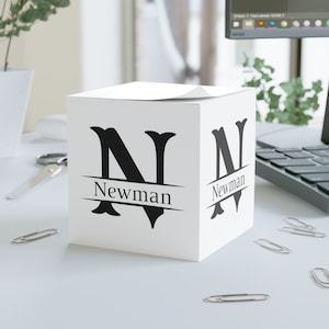 May include: White cube-shaped box with a black letter N and the name Newman printed on each side. The box is sitting on a white surface.