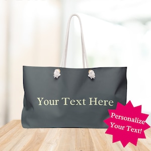 Puede incluir: Una bolsa de lona gris oscuro con asas de cuerda blanca y detalles metálicos dorados. La bolsa tiene el texto "Your Text Here" impreso en color amarillo claro.