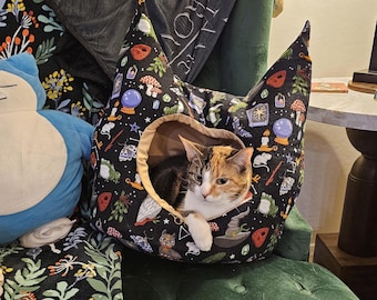 May include: A black cat bed with a whimsical pattern of owls, mushrooms, and other magical elements. The bed has a brown, soft lining and a cat is resting inside.
